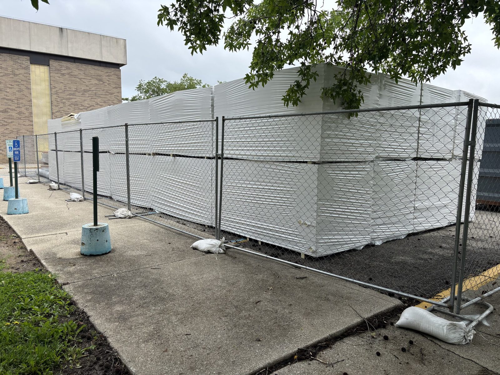 Panel fence on sandbagged stands around wrapped material stacked beside a sidewalk